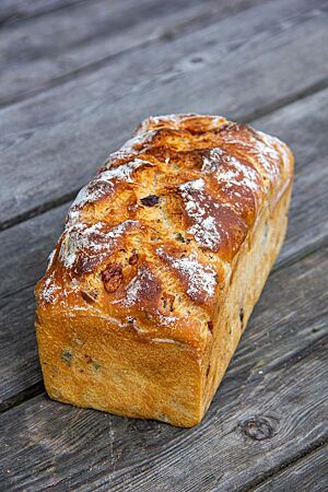Käse-Speck-Oliven-Brot. Das goldbraun ausgebackene, rustikal aufgerissene Käse-Speck-Oliven-Brot mit leicht bemehlter Kruste liegt auf einem Holztisch.