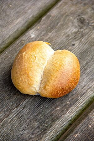 Milchbrötchen (2020). Ein goldgelb ausgebackenes Milchbrötchen mit mittig aufgerissener, glänzender Kruste liegt auf einem rustikalen Holztisch.