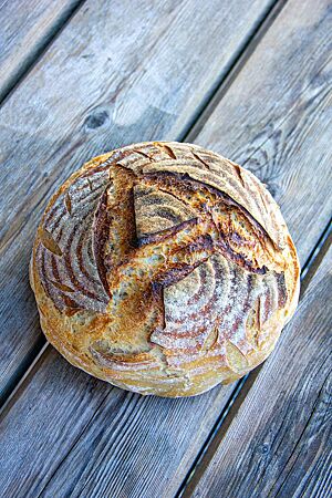 Ein kräftig ausgebackenes Weizensauerteigbrot mit eingeschnittenem „A" auf der Oberfläche liegt auf einem Holztisch.