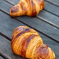 <p>Zwei goldbraun ausgebackene Croissants mit glänzender Oberfläche liegen auf einem rustikalen Holztisch.</p>
