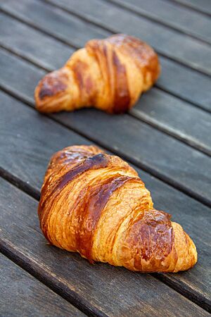<p>Zwei goldbraun ausgebackene Croissants mit glänzender Oberfläche liegen auf einem rustikalen Holztisch.</p>