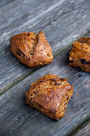 Pflaumen-Walnuss-Ruchmehl-Brötchen. Kräftig ausgebackene, eckig abgestochene und diagonal eingeschnittene Brötchen liegen auf einem rustikalen Holztisch.