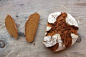 Plan B-Brot (100 % Röstbrot). Das kräftig ausgebackene, rustikal aufgerissene Plan B-Brot mit bemehlter Kruste liegt auf einem Holztisch.