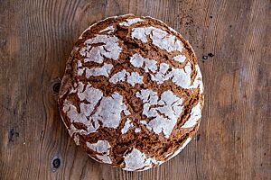 Rauriser Roggenvollkornbrot. Das kräftig ausgebackene Roggenvollkornbrot mit bemehlter Kruste liegt auf einem Holztisch.
