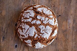 Das kräftig ausgebackene Roggenvollkornbrot mit bemehlter Kruste liegt auf einem Holztisch.