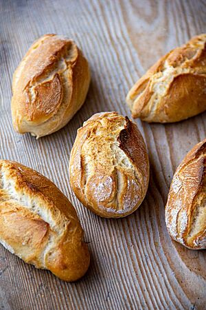 Fünf goldgelb ausgebackene, rustikal aufgerissene Weizenbrötchen mit leicht bemehlter Kruste liegen auf einem Holztisch.