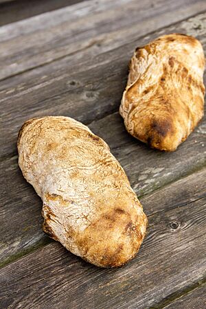 Ciabatta mit kalter Stockgare. Zwei leicht bemehlte Ciabattas mit glatter Oberfläche liegen auf einem rustikalen Holztisch.