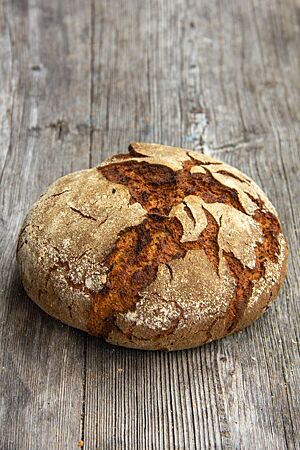 Roggendinkelbrot mit Gewürzen. Das kräftig ausgebackene, rustikal aufgerissene Roggendinkelbrot mit leicht bemehlter Kruste liegt auf einem Holztisch.