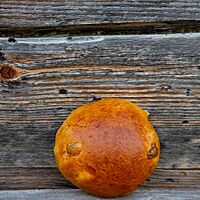 Ein goldbraun ausgebackenes Rosinenbrötchen mit glatter Oberfläche lehnt an einer rustikalen Holzwand.