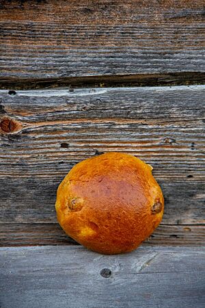 Rosinenbrötchen. Ein goldbraun ausgebackenes Rosinenbrötchen mit glatter Oberfläche lehnt an einer rustikalen Holzwand.
