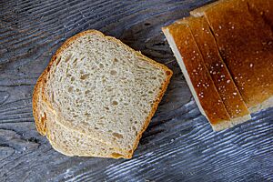 Zwei Scheiben Sandwichbrot liegen neben dem goldbraun ausgebackenen Laib mit matter Oberfläche und etwas Schrot auf der Kruste.