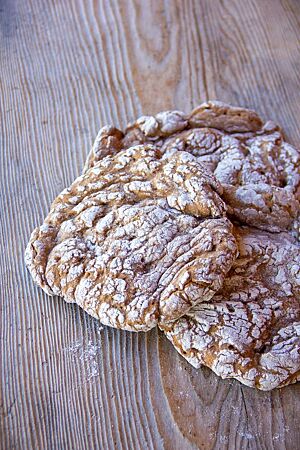 Schüttelbrot (2017). Drei kleine, flache Brote mit Rissen auf der bemehlten Oberfläche liegen auf einem Holztisch.