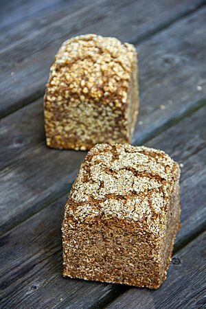 Schwarzbrot mit Sonnenblumenkernen. Zwei ausgebackene Kastenbrote mit Schrot bzw. Haferflocken auf der rissigen Kruste liegen auf einem rustikalen Holztisch.