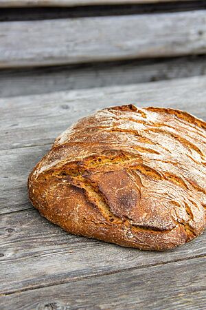 Schwarzweißbrot. Ein kräftig ausgebackener, recht flacher Brotlaib mit leicht bemehlter Kruste liegt auf einem rustikalen Holztisch.