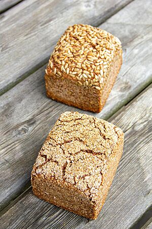 Zwei kräftig ausgebackene Schrotbrote liegen auf einem rustikalen Holztisch: Das hintere ist mit Sonnenblumenkernen bestreut, das vordere mit Schrot.