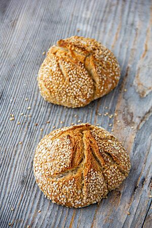 Sesambrötchen. Zwei goldbraun ausgebackene, rustikal aufgerissene Weizenbrötchen mit Sesam auf der Kruste liegen auf einem Holztisch.