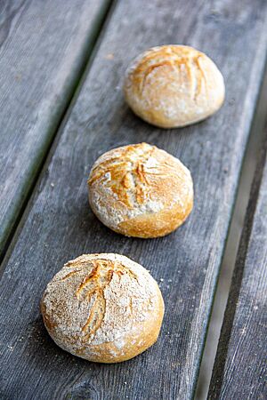 Sonntagsbrötchen. Drei goldgelb ausgebackene Sonntagsbrötchen mit bemehlter und wild aufgerissener Kruste liegen in einer Reihe hintereinander auf einer rustikalen Holzbank.