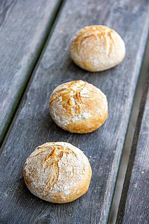 Drei goldgelb ausgebackene Sonntagsbrötchen mit bemehlter und wild aufgerissener Kruste liegen in einer Reihe hintereinander auf einer rustikalen Holzbank.