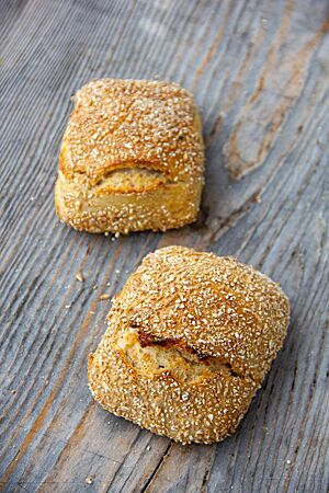 Sonntagsdinkelbrötchen. Zwei goldgelb ausgebackene Sonntagsdinkelbrötchen mit Schrot auf der Kruste liegen auf einem rustikalen Holztisch.