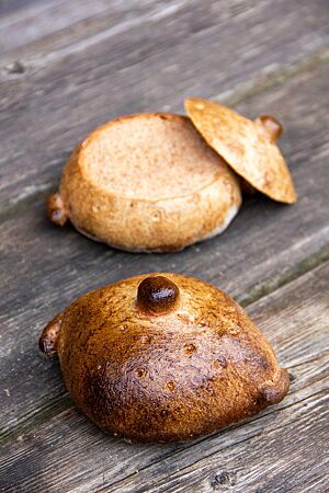 Tassenbrot. Zwei kleine, goldbraun ausgebackene Brote in Form von Suppentassen liegen auf einer rustikalen Holzbank. Oben und seitlich der Brote sind gebackene Teigstücke in Form von Deckelknopf und Henkel angebracht. Das abgeschnittene obere Drittel des hinteren Brotes symbolisiert den Deckel der Suppentasse.
