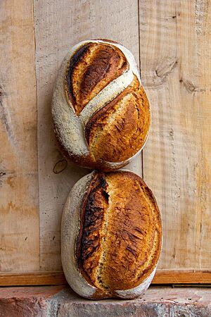 Zwei rustikal aufgerissene Brotlaibe stehen auf einem Ziegelstein, lehnend an einer Holzwand. Das obere ist zweimal spitzwinklig zur Längsachse eingeschnitten, das untere der Länge nach mittig.