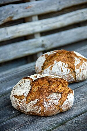 Thomas’ Weizenlaib (30-Kräuter-Brot). Zwei kräftig ausgebackene Weizenlaibe liegen auf einer rustikalen Holzbank.
