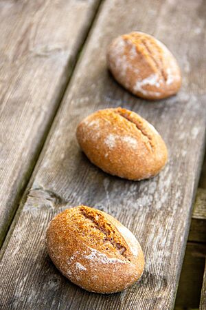 Die rustikal aufgerissenen Vollkornbrötchen mit leicht bemehlter Kruste liegen auf einem Holztisch.