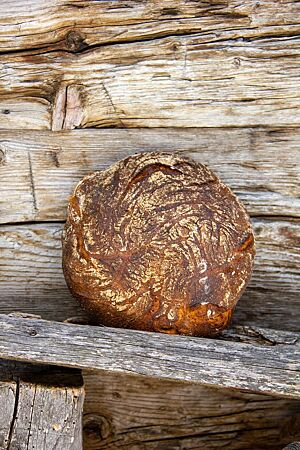 Das kräftig ausgebackene Wiesenkräuterbrot mit leicht bemehlter Kruste lehnt an einer rustikalen Holzwand.