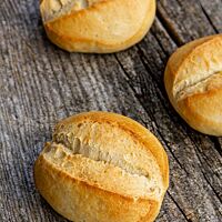 Drei ovale Brötchen mit mittig eingeschnittener Kruste liegen auf einem rustikalen Holztisch.