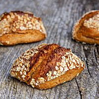 Drei kräftig ausgebackene Dinkelvollkornbrötchen mit Haferflocken auf der Oberfläche liegen auf einem rustikalen Holztisch.