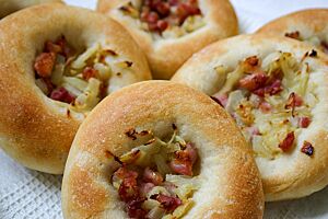 Bialys. Die goldgelb ausgebackenen Brötchen sind in der Mitte mit gedünstetem Bauchspeck, Zwiebeln und Knoblauch belegt und haben einen breiten Rand.