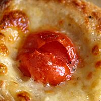 Das Kartoffel-Pizza-Nest ist ein rundes Gebäckstück, das mit Mozzarella überbacken ist und in dessen Mitte eine kleine, tief eingedrückte Tomate liegt.