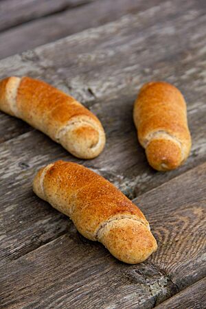 Rohlíky graham. Drei längliche, zu Stangen aufgerollte Brötchen liegen auf einem rustikalen Holztisch.