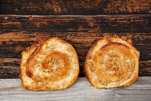 Vollkornpuddingschnecken. Zwei mit Streuseln belegte Vollkornpuddingschnecken lehnen an einer rustikalen Holzwand.