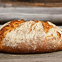 Das kräftig ausgebackene Willkommensbrot mit bemehlter Kruste liegt auf einem rustikalen Holztisch.