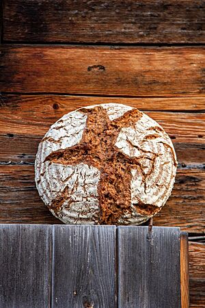 Ein kräftig ausgebackenes, rustikal aufgerissenes Vollkornbrot mit bemehlter Kruste lehnt an einer Holzwand.