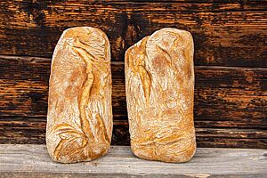 Zwei goldgelb ausgebackene Ciabattas mit leicht bemehlter und etwas aufgerissener Kruste lehnen an einer Holzwand.