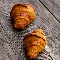Zwei goldgelb ausgebackene Dinkelcroissants mit glänzender Oberfläche liegen auf einem rustikalen Holztisch.