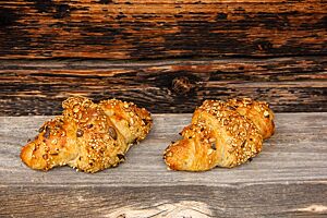 Saatencroissants. Zwei hellbraun ausgebackene Croissants mit einer Saatenmischung auf der Kruste liegen auf einem rustikalen Holztisch.