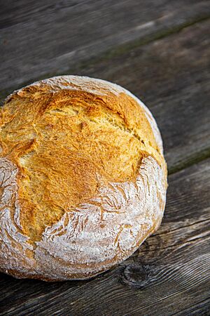 Ein goldgelb ausgebackenes, rustikal aufgerissenes Weizenbrot mit leicht bemehlter Kruste liegt auf einem Holztisch.