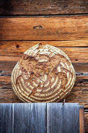 Sarahs Sauerteigbrot mit Einkorn und Leinsaat. Ein kräftig ausgebackenes, rustikal aufgerissenes Sauerteigbrot mit leicht bemehlter Kruste lehnt an einer Holzwand.