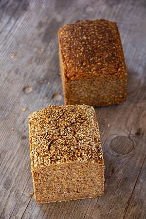 Ganzkorn-Roggenschrotbrot. Zwei kräftig ausgebackene Kastenbrote, eines davon mit Schrot auf der Kruste, liegen auf einem grauen Holztisch.
