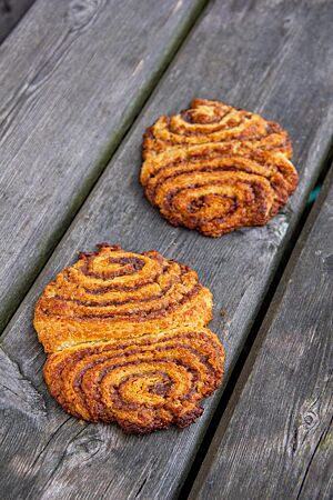 Einkorn-Franzbrötchen. Zwei Franzbrötchen mit goldgelber Farbe und knuspriger, karamellisierter Zimtfüllung liegen auf einem grauen Holztisch.
