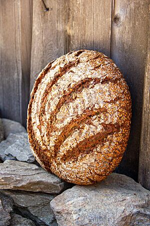 Ein kräftig ausgebackener, auf der Oberfläche eingeschnittener Brotlaib mit bemehlter Kruste lehnt an einer rustikalen Holzwand.