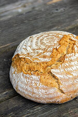 Dinkelkrustenbrot. Ein runder, goldgelb ausgebackener Laib mit rustikalem Ausbund und leicht bemehlter Kruste liegt auf einem dunkelgrauen Holztisch.