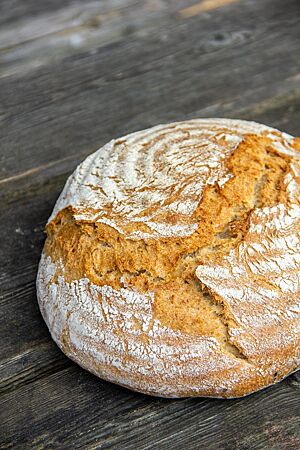 Ein runder, goldgelb ausgebackener Laib mit rustikalem Ausbund und leicht bemehlter Kruste liegt auf einem dunkelgrauen Holztisch.