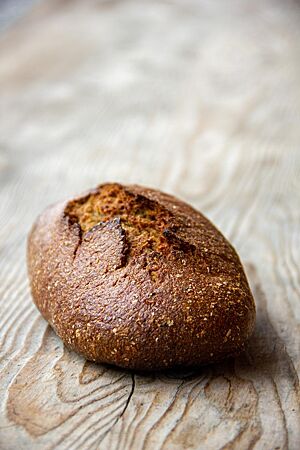 Ein dunkelbraun ausgebackenes, rustikal aufgerissenes Roggenmischbrot mit glänzender Oberfläche liegt auf einem Holztisch.