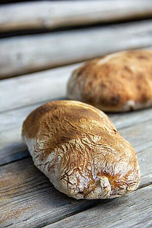 Hüttenpantoffel (Ciabatta). Ein kräftig ausgebackenes Ciabatta mit leicht bemehlter und stellenweise dunkelbrauner Kruste liegt auf einem Holztisch.