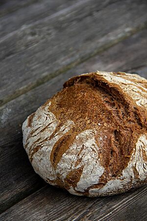Rotkornweizen-Sauerteigbrot. Ein kräftig ausgebackenes, rustikal aufgerissenes Vollkornbrot mit bemehlter Kruste liegt auf einem Holztisch.