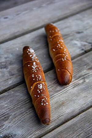 Almstangerl. Zwei dunkel ausgebackene, mit grobem Salz bestreute Almstangerl liegen auf einem rustikalen Holztisch.